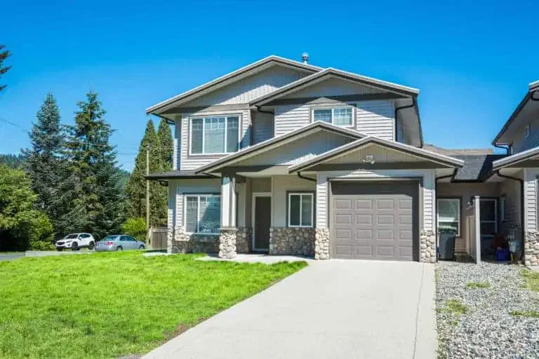 A modern two-story house with a gray exterior and stone accents, featuring a garage and a neatly paved driveway, located in a sunny suburban area.