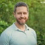 A man with short gray hair and a beard smiles at the camera, standing outdoors in front of green foliage, wearing a light blue polo shirt.