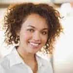 Person with curly hair smiling and wearing a white shirt, in a well-lit setting.