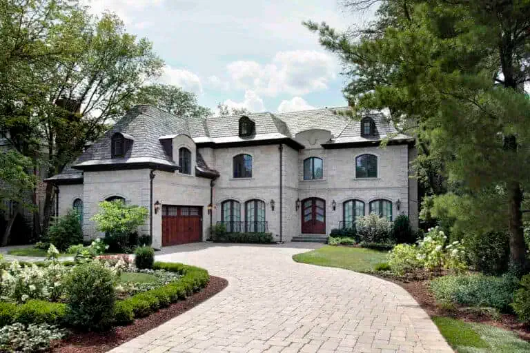 A large stone house with a paved driveway, multiple arched windows, wooden garage doors, and a landscaped front yard with bushes and trees.