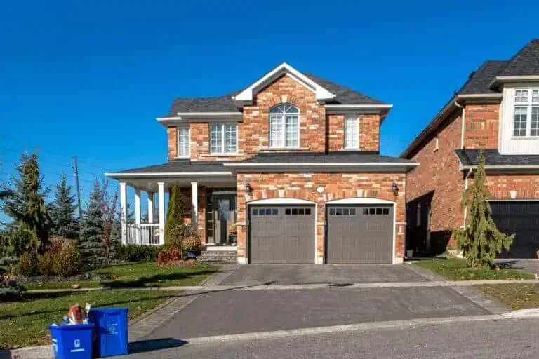 A two-story brick house with a double garage, driveway, and a front porch is seen on a clear day. A blue recycling bin and a brown paper bag are placed at the end of the driveway.