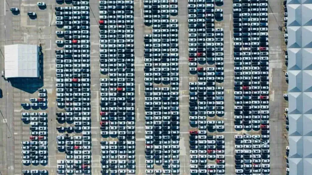 Aerial view of a large parking lot filled with rows of cars, organized in a grid pattern. A white canopy structure is located on the left side.