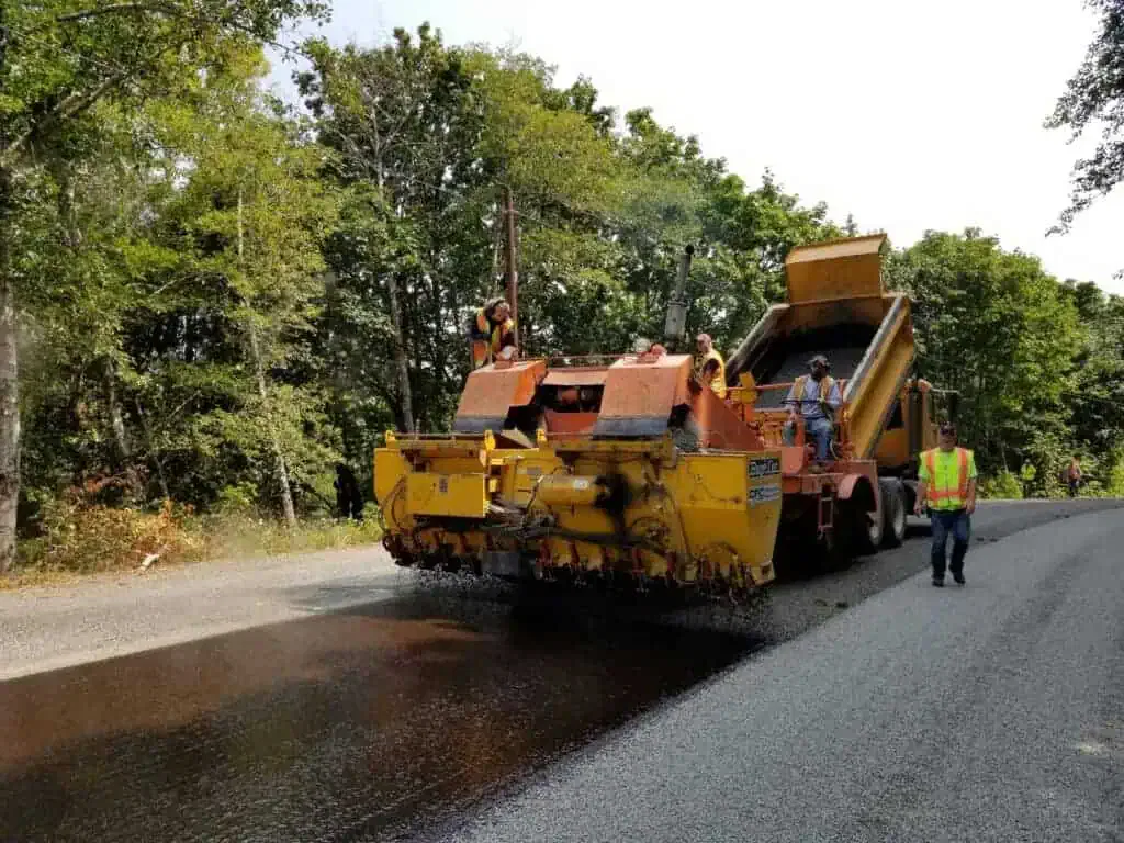Road construction workers operate machinery to lay asphalt on a rural road surrounded by trees. A worker in a safety vest walks nearby.