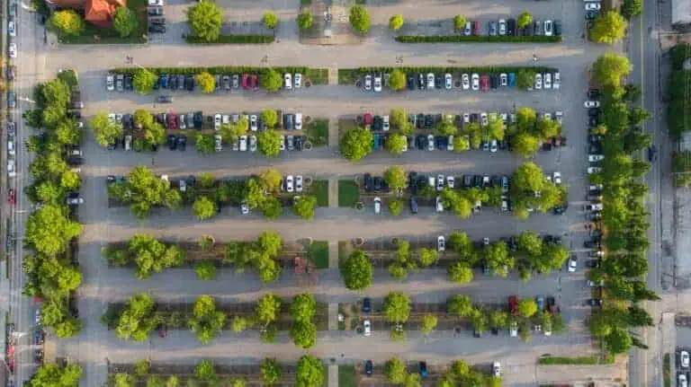 Aerial view of a large parking lot filled with parked cars, featuring multiple rows separated by lines of trees and greenery.