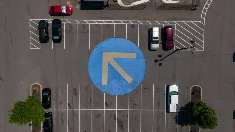 Aerial view of a parking lot with several cars parked in designated spots. A large blue circle with a white arrow pointing up and left is marked on the ground in the center of the lot.