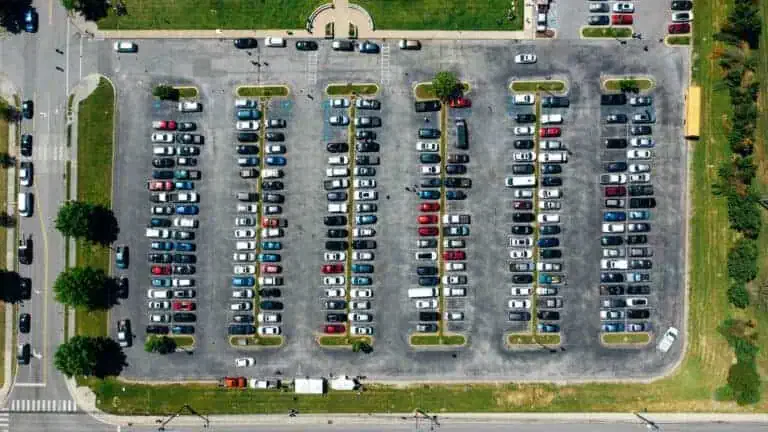 Aerial view of a large parking lot filled with rows of parked cars and a yellow school bus near the upper right corner, surrounded by green grass and trees.