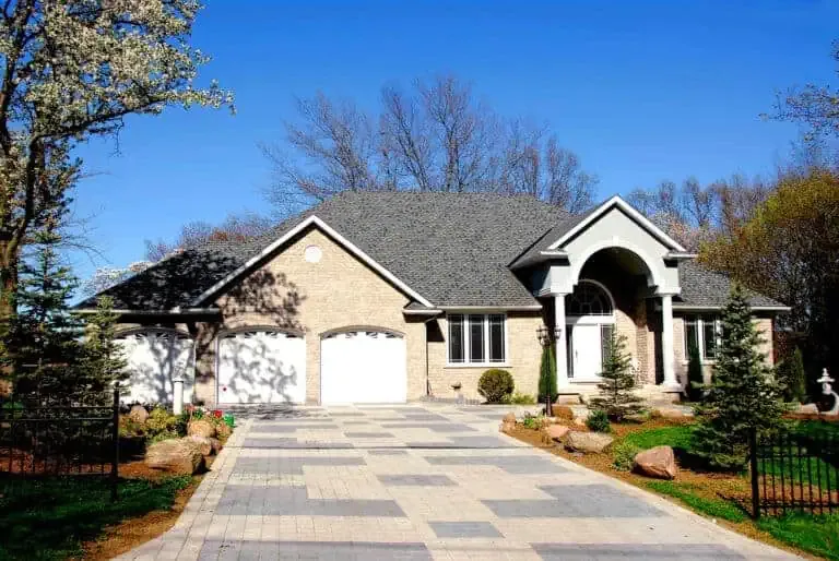 A single-story suburban home with a large arched entryway, stone facade, and double garage doors, surrounded by blooming trees under a clear blue sky.