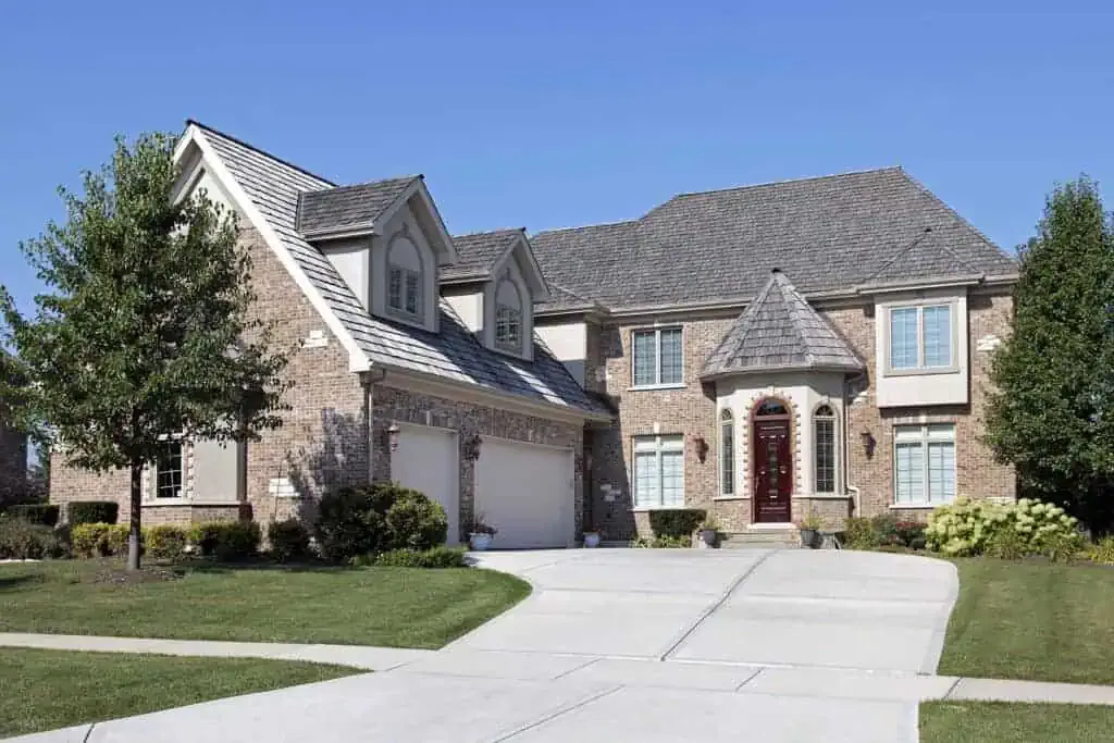 Large two-story suburban house with a stone and brick facade, gabled roofs, a double garage, and a neatly maintained front lawn under a clear blue sky.