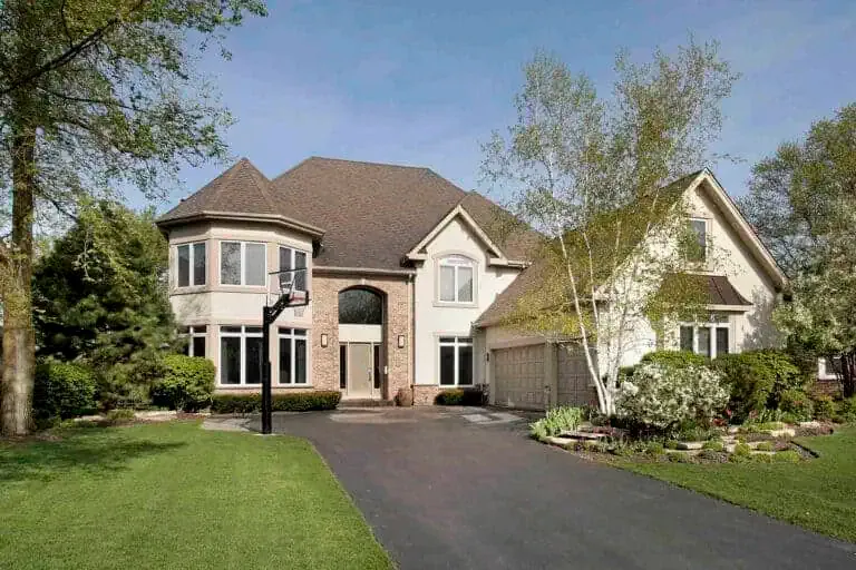 A two-story house with beige siding, a brick entryway, and a brown roof. It has a three-car garage, manicured landscaping, and a basketball hoop in the driveway.