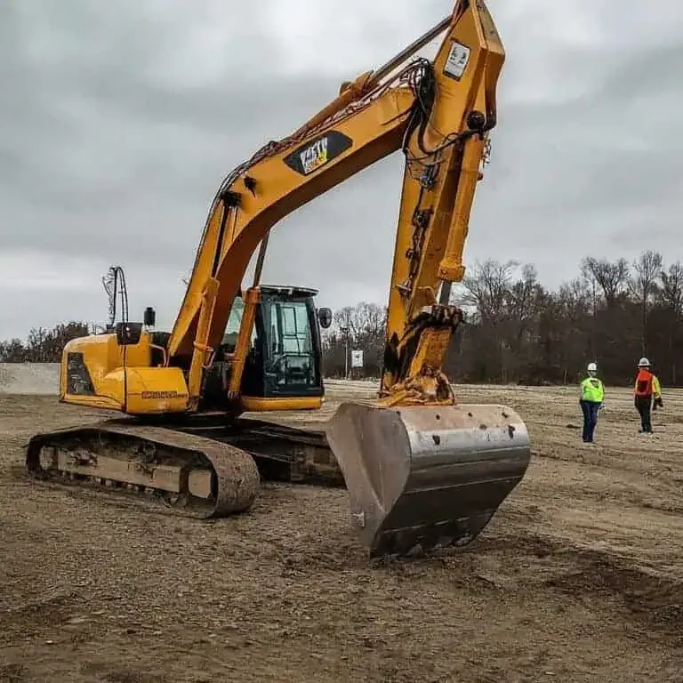 A yellow excavator is parked on a construction site with two workers wearing safety gear walking nearby.