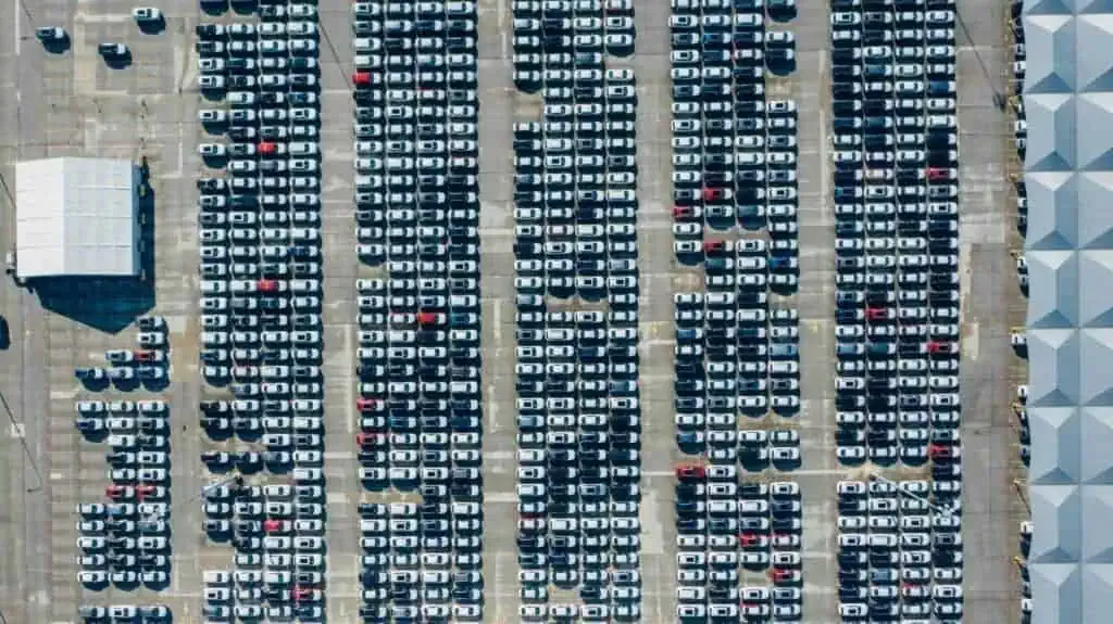 Aerial view of a large parking lot filled with rows of cars, organized in a grid pattern. A white canopy structure is located on the left side.