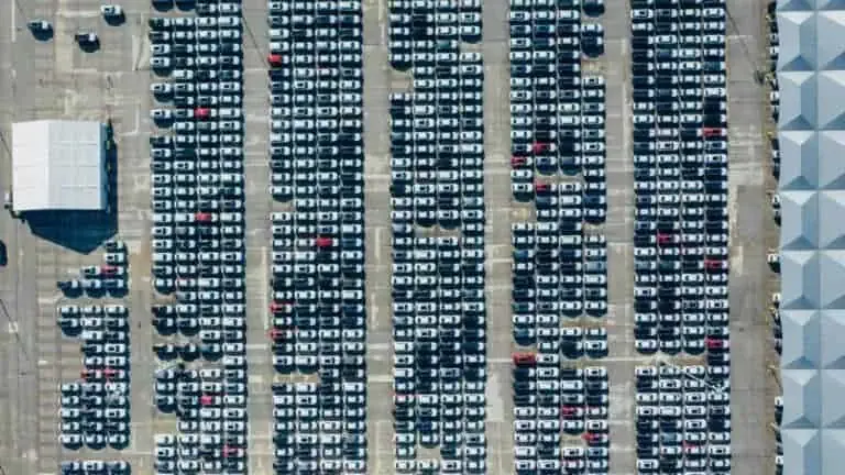 Aerial view of a large parking lot filled with rows of cars, organized in a grid pattern. A white canopy structure is located on the left side.