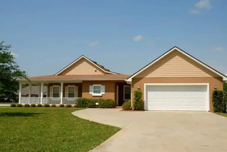 Single-story tan house with a two-car garage, front porch, and well-maintained lawn under a clear blue sky.