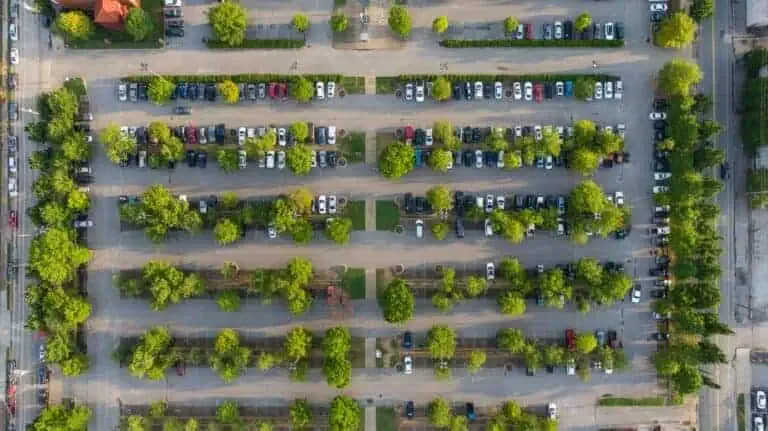 Aerial view of a large parking lot filled with parked cars, featuring multiple rows separated by lines of trees and greenery.