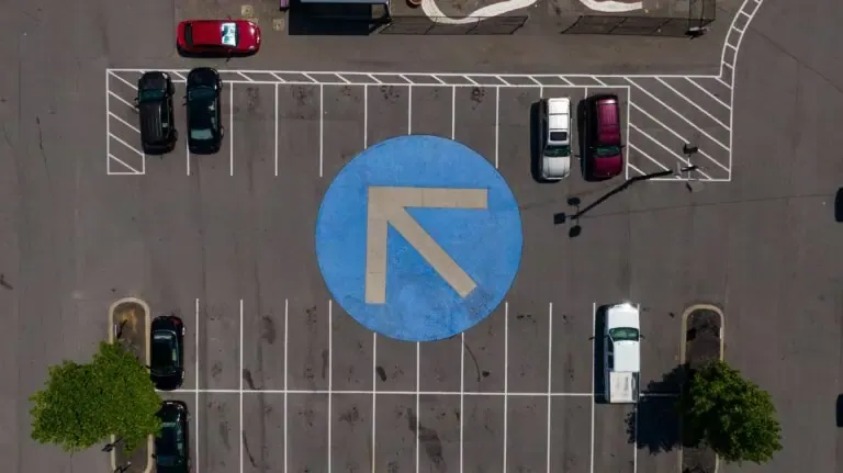 Aerial view of a parking lot with several cars parked in designated spots. A large blue circle with a white arrow pointing up and left is marked on the ground in the center of the lot.