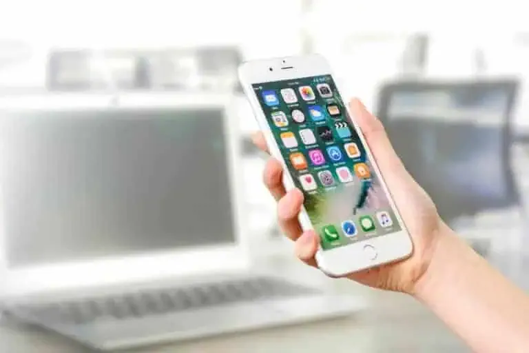 A hand holding a white smartphone displaying various app icons, with a blurred open laptop in the background.